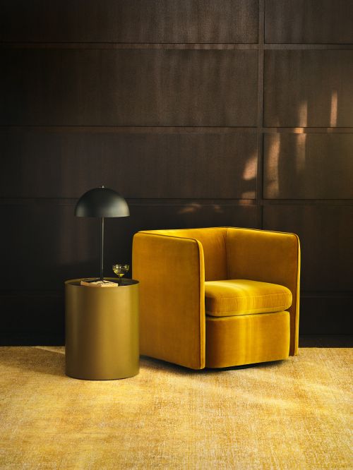 Interior design featuring a mustard-yellow velvet chair and a side table with a lamp against a dark panelled wall and matching carpet.