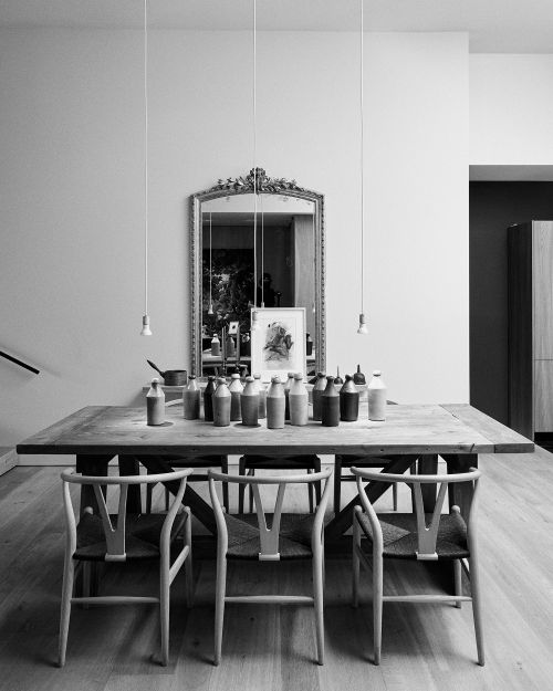 Monochrome photo of a wooden dining table with wishbone chairs surrounded by an arrangement of bottles in an indoor setting.