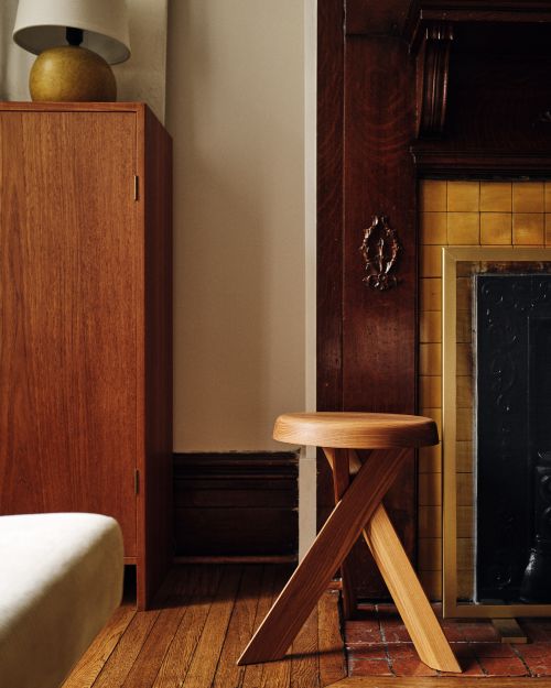 Wooden stool with a round seat and criss-cross legs next to a wooden cabinet and fireplace in a neutral-toned room.