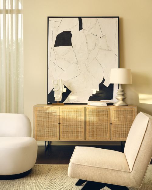 Living room interior with tan sideboard, abstract art, table lamp, and seating. Warm lighting and neutral color palette.