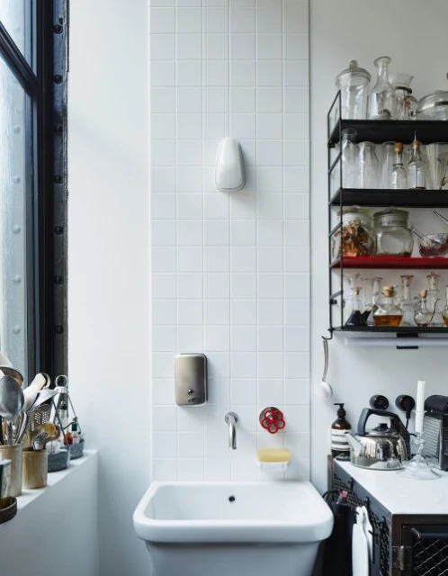 Clean white bathroom interior with a white sink, wall-mounted soap dispensers, tiled walls, and window. Shelving with glass jars.