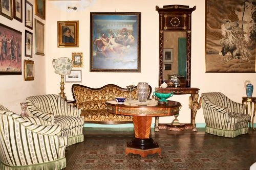 Ornate sitting room featuring striped armchairs, patterned sofa, round table, and various framed artworks on the pale walls.
