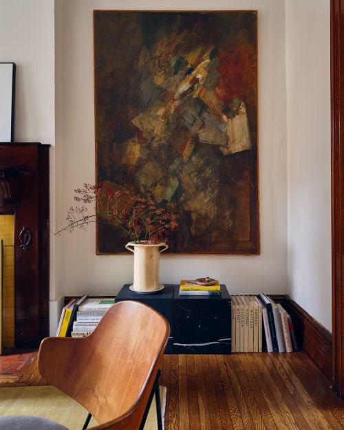 Interior shot featuring a wooden chair, a large dark abstract painting, a ceramic vase with dried branches, and stacked books.