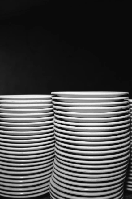 Stack of many white ceramic plates, arranged in two main piles against a dark background, showcasing repetition and clean lines.