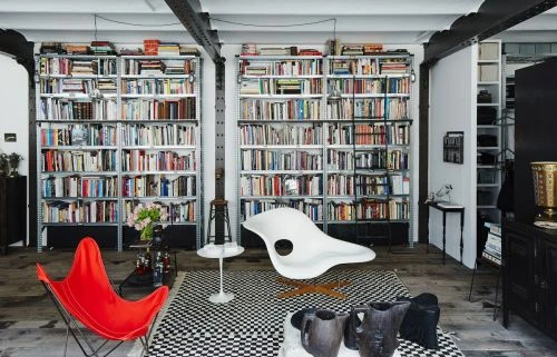 Interior view of a modern living room featuring bookshelves, a red butterfly chair, a white chair, black decorative vases, and a black and white rug.