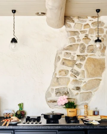 Indoor kitchen scene featuring a black stove top with a pot, a Ricard mug, groceries, flowers, a stone wall, and two hanging lights.