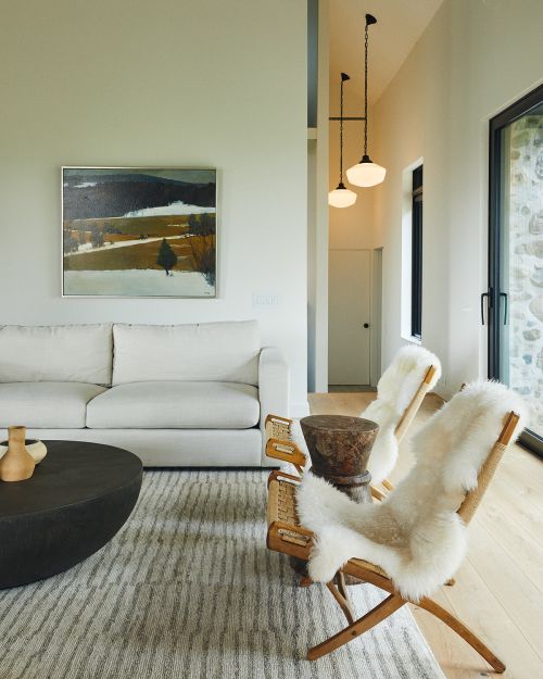 Interior view of a modern living room with a white sofa, round coffee table, two wooden chairs draped with sheepskins, and landscape painting.