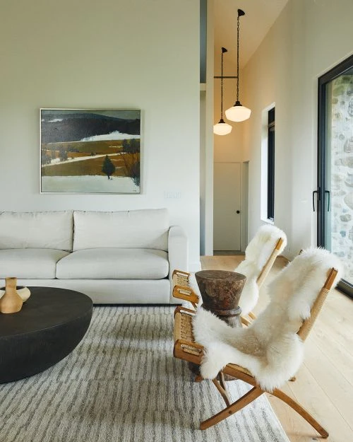 Interior view of a modern living room with a white sofa, round coffee table, two wooden chairs draped with sheepskins, and landscape painting.