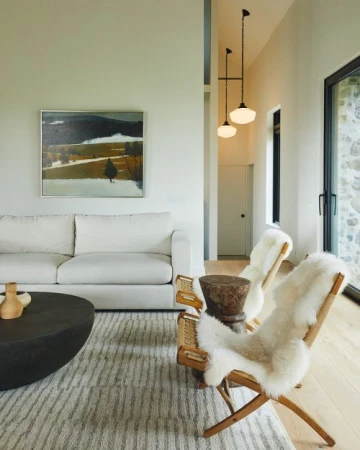 Interior view of a modern living room with a white sofa, round coffee table, two wooden chairs draped with sheepskins, and landscape painting.