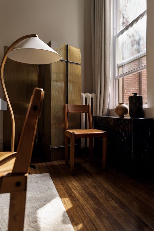 Interior view of room with chair, floor lamp, cabinet, marble-topped furniture piece, wooden floor and window with curtains.