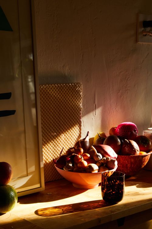 Warm-toned still life featuring assorted fruit in bowls with a dark glass on a wooden surface in bright, natural lighting indoors.