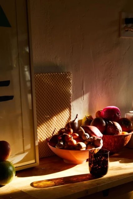 Warm-toned still life featuring assorted fruit in bowls with a dark glass on a wooden surface in bright, natural lighting indoors.