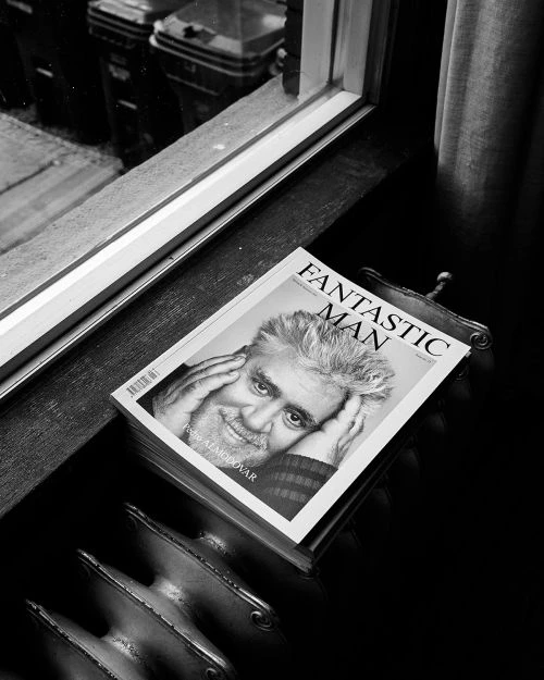 Black and white photo of a stack of Fantastic Man magazines by a window, featuring Pedro Almodóvar on the cover, with a metal radiator visible.