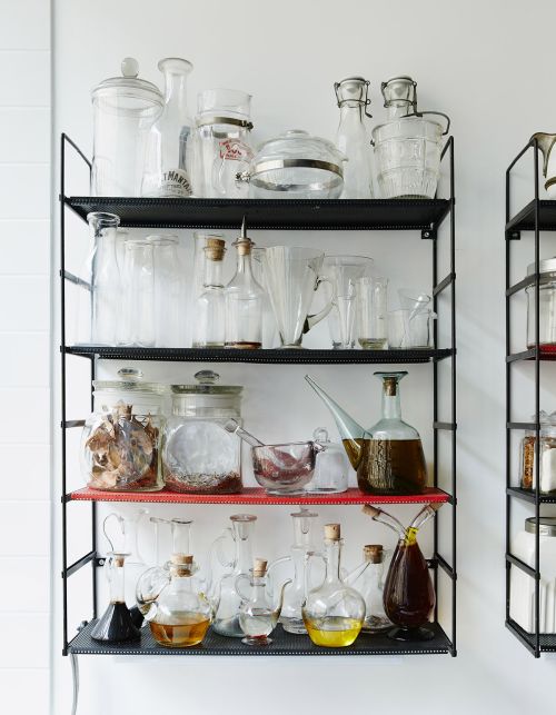 A collection of various clear glass jars and bottles displayed on a multi-tiered black metal shelf against a white wall, clean aesthetic.