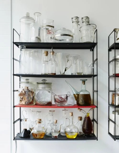 A collection of various clear glass jars and bottles displayed on a multi-tiered black metal shelf against a white wall, clean aesthetic.
