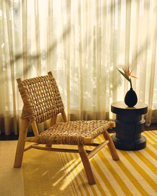 Woven chair with wood frame on a striped rug, next to a small table with a vase and a flower, in front of sheer curtains.