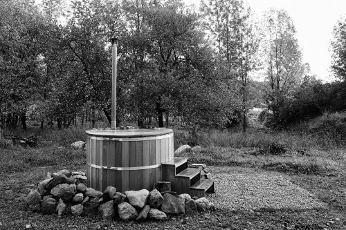 Outdoor wooden hot tub with a metal chimney, surrounded by rocks and gravel, set against a backdrop of trees in a rural landscape.