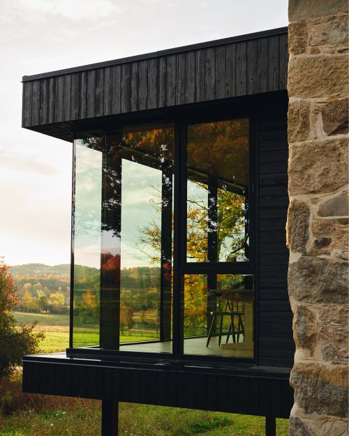Exterior view of a modern house with black wood siding and large windows, reflecting the surrounding landscape of trees and fields.