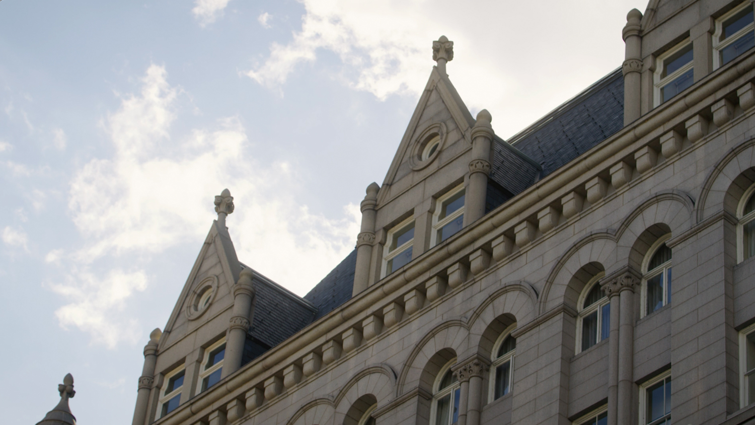 Exterior of a grey stone building with peaked roof, multiple windows, and decorative architectural details against a cloudy sky.