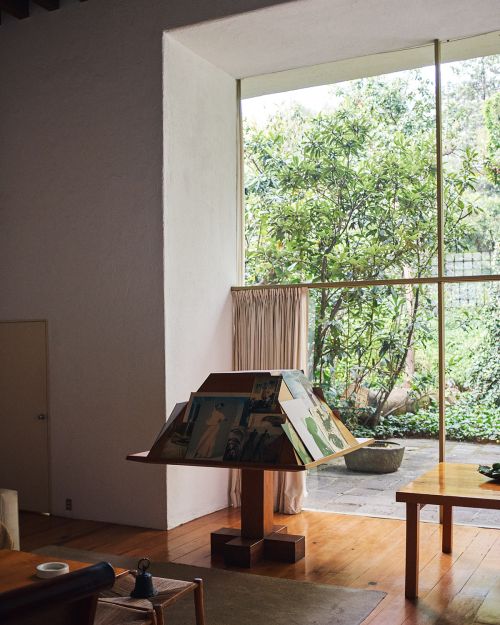 Brightly lit interior shot of a room with hardwood floors, a geometric table displaying books, and a large window overlooking a leafy garden.