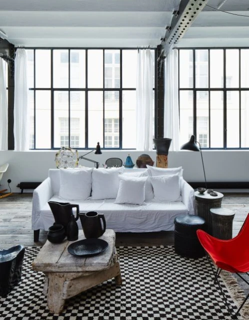 Living room interior features a white sofa, black-and-white checkered rug, and natural light from large windows with white curtains.