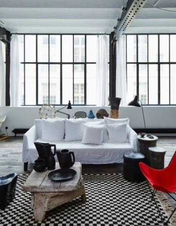 Living room interior features a white sofa, black-and-white checkered rug, and natural light from large windows with white curtains.