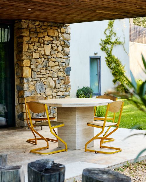 Octagonal stone dining table surrounded by yellow-framed chairs on a patio with a stone pillar and modern building exterior