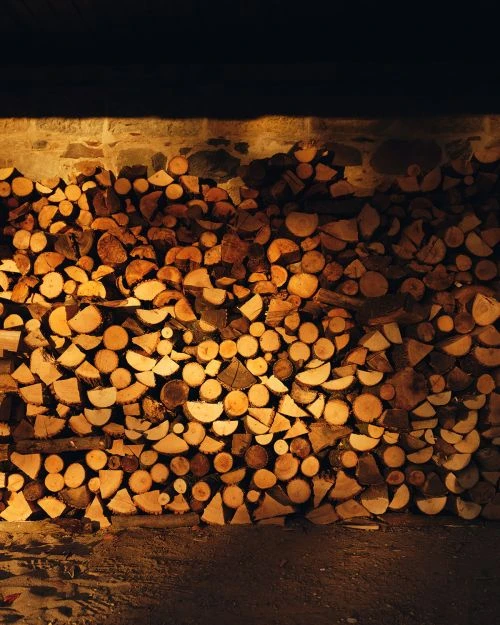 Stack of chopped firewood stacked against a stone wall, in a low-lit, indoor setting, showing rustic textures and warm, earthy tones.