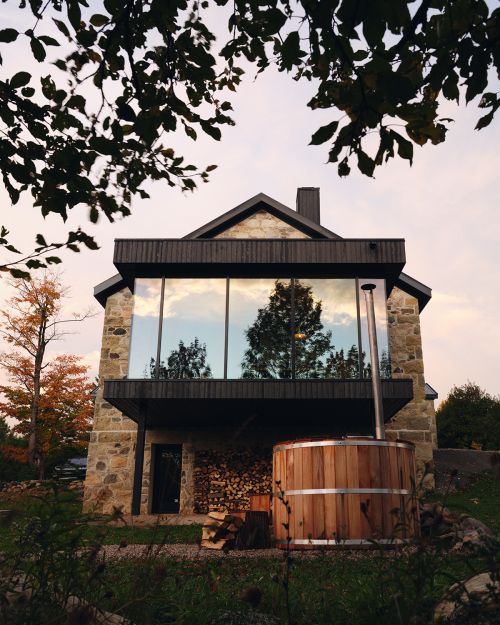 Exterior of stone building with a balcony, large windows reflecting trees, and a wooden hot tub in the foreground at dusk.
