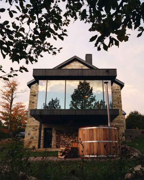 Exterior of stone building with a balcony, large windows reflecting trees, and a wooden hot tub in the foreground at dusk.