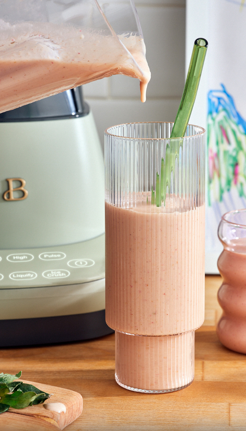 Peach smoothie is poured from a blender into a glass with a green straw on a wooden counter with a child's drawing in the background.