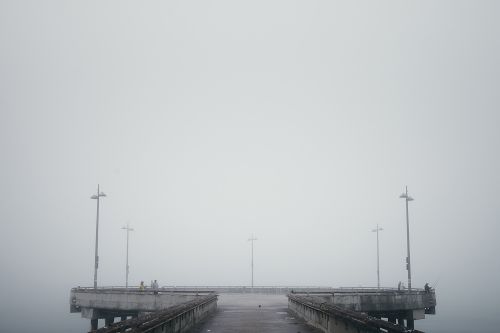 Foggy day at the pier featuring silhouettes of people and lampposts. The end of the pier extends into the misty horizon.