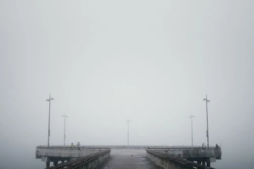 Foggy day at the pier featuring silhouettes of people and lampposts. The end of the pier extends into the misty horizon.