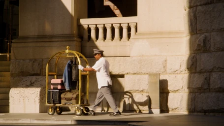 Hotel bellhop in white uniform pushing a brass luggage cart loaded with bags in front of a stone building.