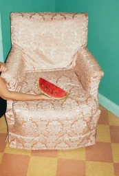 An armchair upholstered in patterned damask fabric holding a single slice of watermelon on the seat cushion.