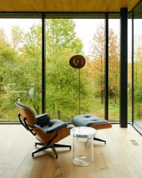 Modern lounge chair and ottoman positioned by a large floor-to-ceiling glass wall overlooking autumn trees.