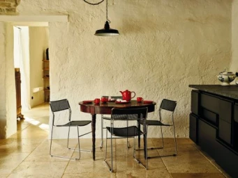 Round wooden dining table with red teapot and cups surrounded by four metal wire chairs against a textured plaster wall