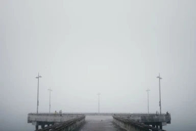 Wide horizontal photograph of a concrete pier extending into thick fog with distant light poles