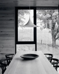Black and white interior photograph of a modern dining table with a ceramic bowl beneath a pendant lamp, overlooking trees.