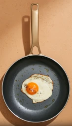 Overhead shot of a single fried egg cooking in a frying pan with a golden handle on a textured beige surface