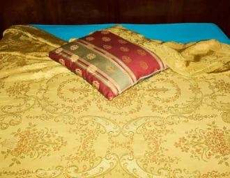 Close-up of a bed with patterned gold damask bedspread, bright turquoise sheet, and a red decorative pillow.