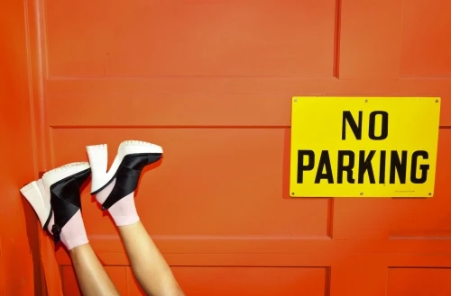 Upside-down view of woman's legs with block-heel boots, pale pink socks, against an orange door with a yellow 'No Parking' sign.