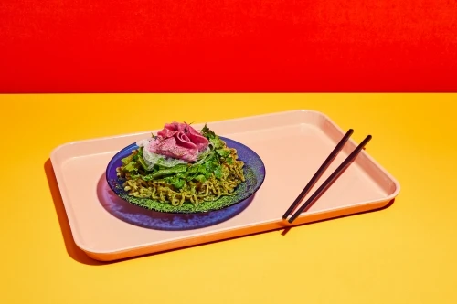 Overhead shot of vibrant green noodles with meat and garnish on a blue plate with chopsticks and pink tray against red and yellow backdrop.