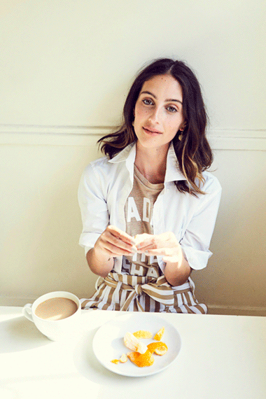 Young woman in a casual outfit sits at a white table, peeling fruit next to a cup of coffee in a brightly lit indoor setting.