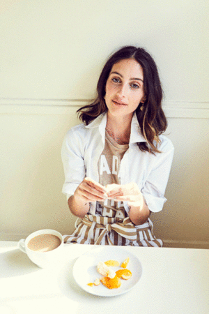 Young woman in a casual outfit sits at a white table, peeling fruit next to a cup of coffee in a brightly lit indoor setting.