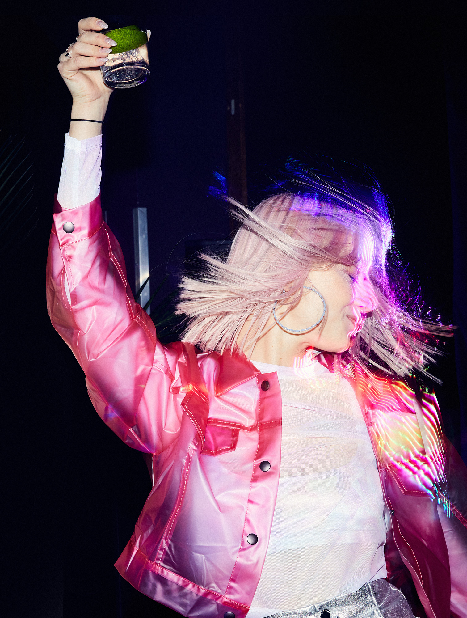 Woman with pink hair in a pink jacket holds up a cocktail glass in an indoor setting, celebrating or enjoying the drink.