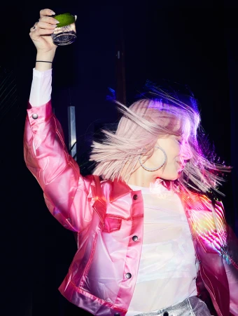Woman with pink hair in a pink jacket holds up a cocktail glass in an indoor setting, celebrating or enjoying the drink.