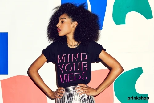 A woman with dark, curly hair poses against a colorful, geometric backdrop, wearing a black t-shirt and silver skirt for a fashion shot.