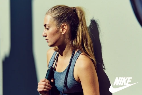 A blonde female athlete in sportswear holds a jump rope against a white wall, with shadows and the Nike logo visible.