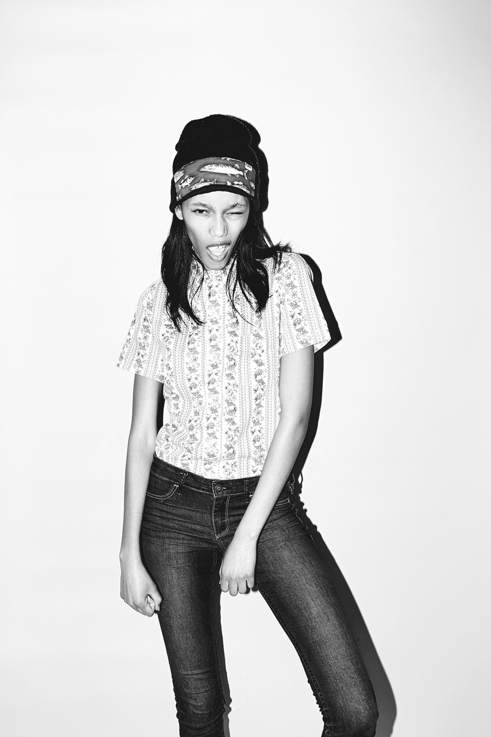 Young woman wearing a beanie and patterned t-shirt posing playfully against a white wall